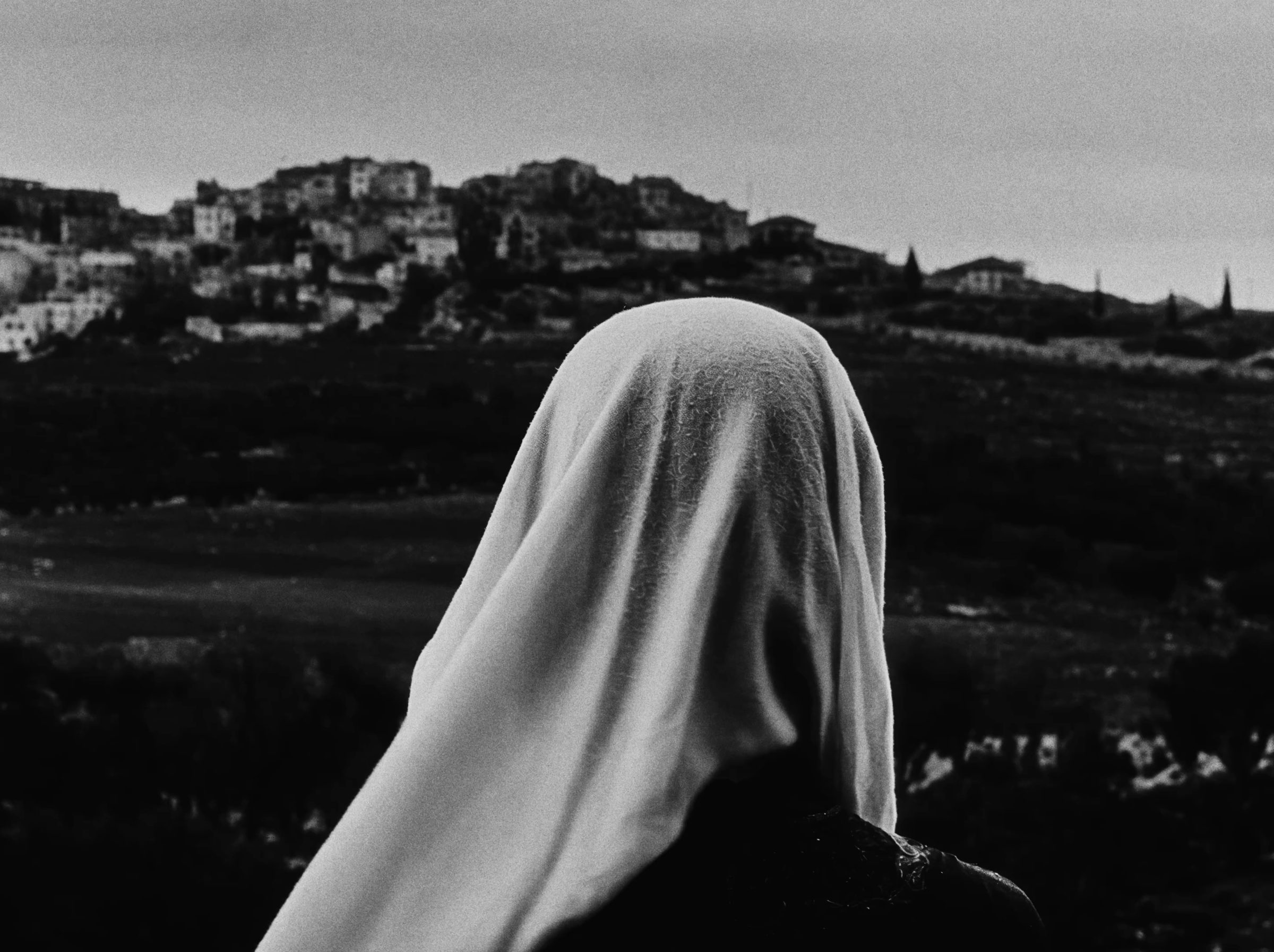 A veiled woman, seen from behind, looks toward a village on a hill.